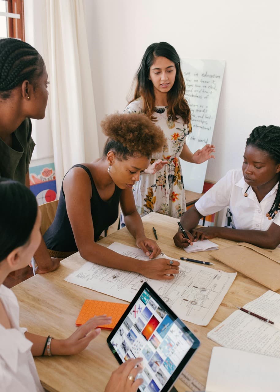 A team of 5 women are gathered around a blueprint strategizing with a tablet in hand